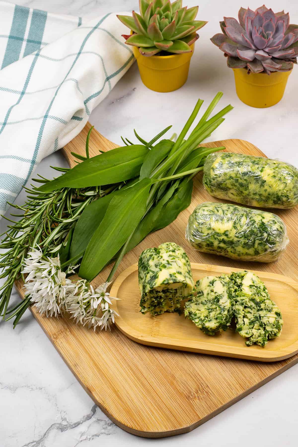 3 logs of wild garlic and rosemary butter on a board with sprigs of rosemary and leaves and flowers of wild garlic on the side.