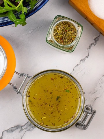 Zesty Italian-style dressing in a jar with a bowl of mixed leaf salad and jar of Italian seasoning on the side.