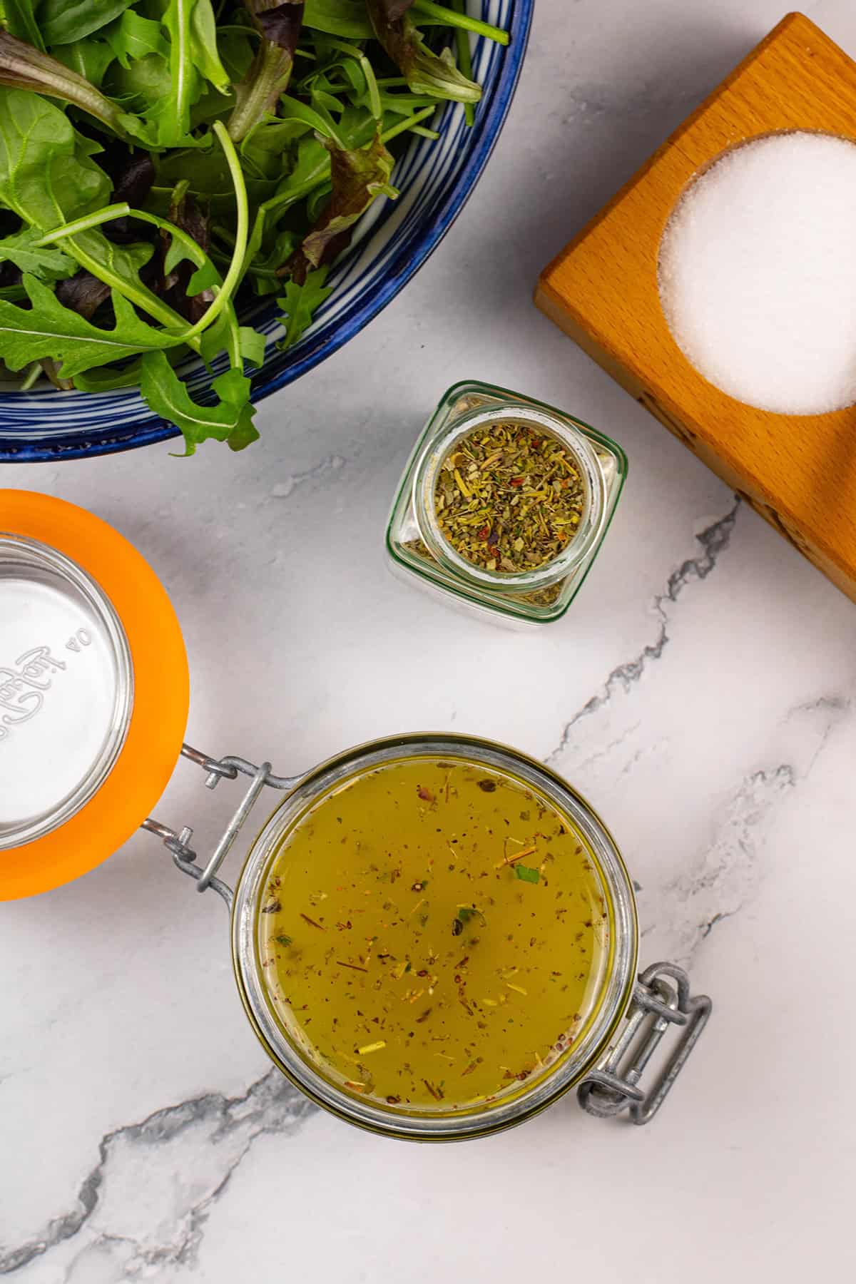 Zesty Italian-style dressing in a jar with a bowl of mixed leaf salad and jar of Italian seasoning on the side.