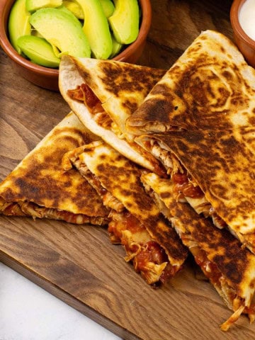 Chicken tinga quesadillas on a board with a bowl of salsa and dishes of sliced avocado and sour cream.