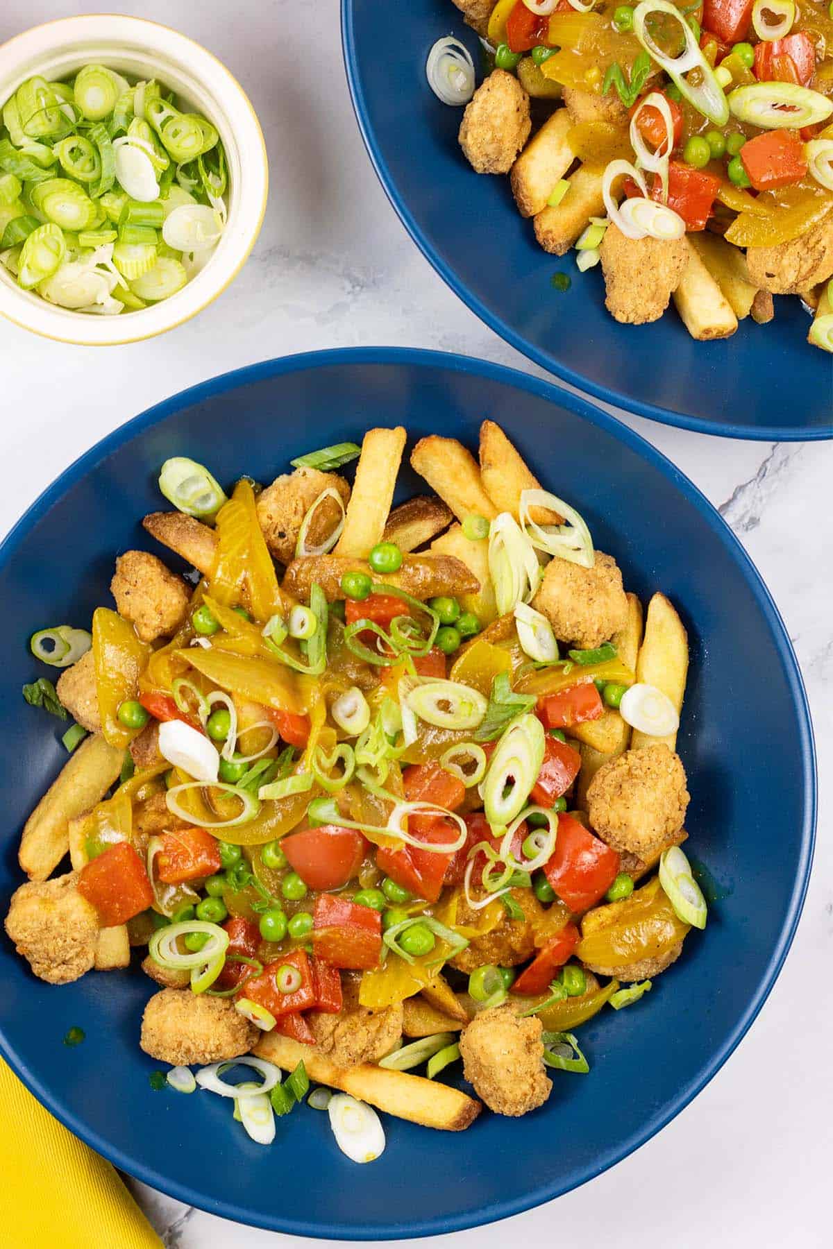 2 portions of crispy chicken curry loaded fries in dark blue bowls with a small dish of sliced spring onions and a yellow napkin on the side.