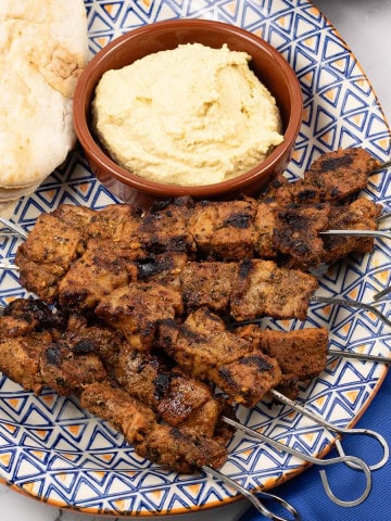 Easy greek pork skewers on an orange blue and white patterned platter with pitta breads and houmous.