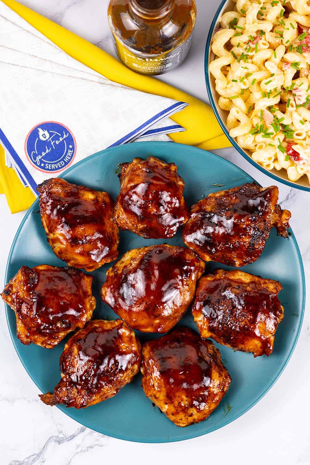 Juicy BBQ chicken thighs on a teal plate with a bottle of barbecue sauce and bowl of pasta salad on the side.