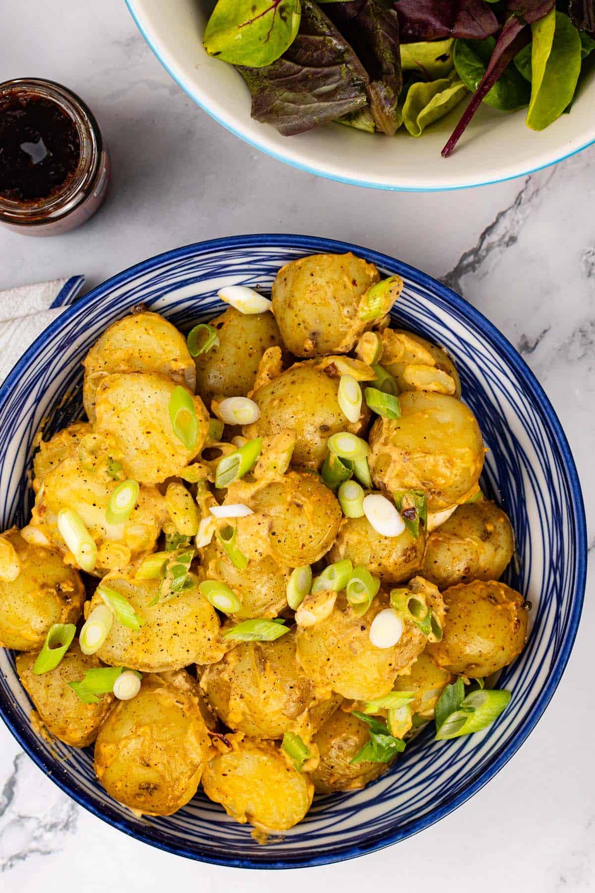 Smoky chipotle potato salad in a blue and white serving bowl, with a jar of chipotle paste and bowl of salad leaves on the side.