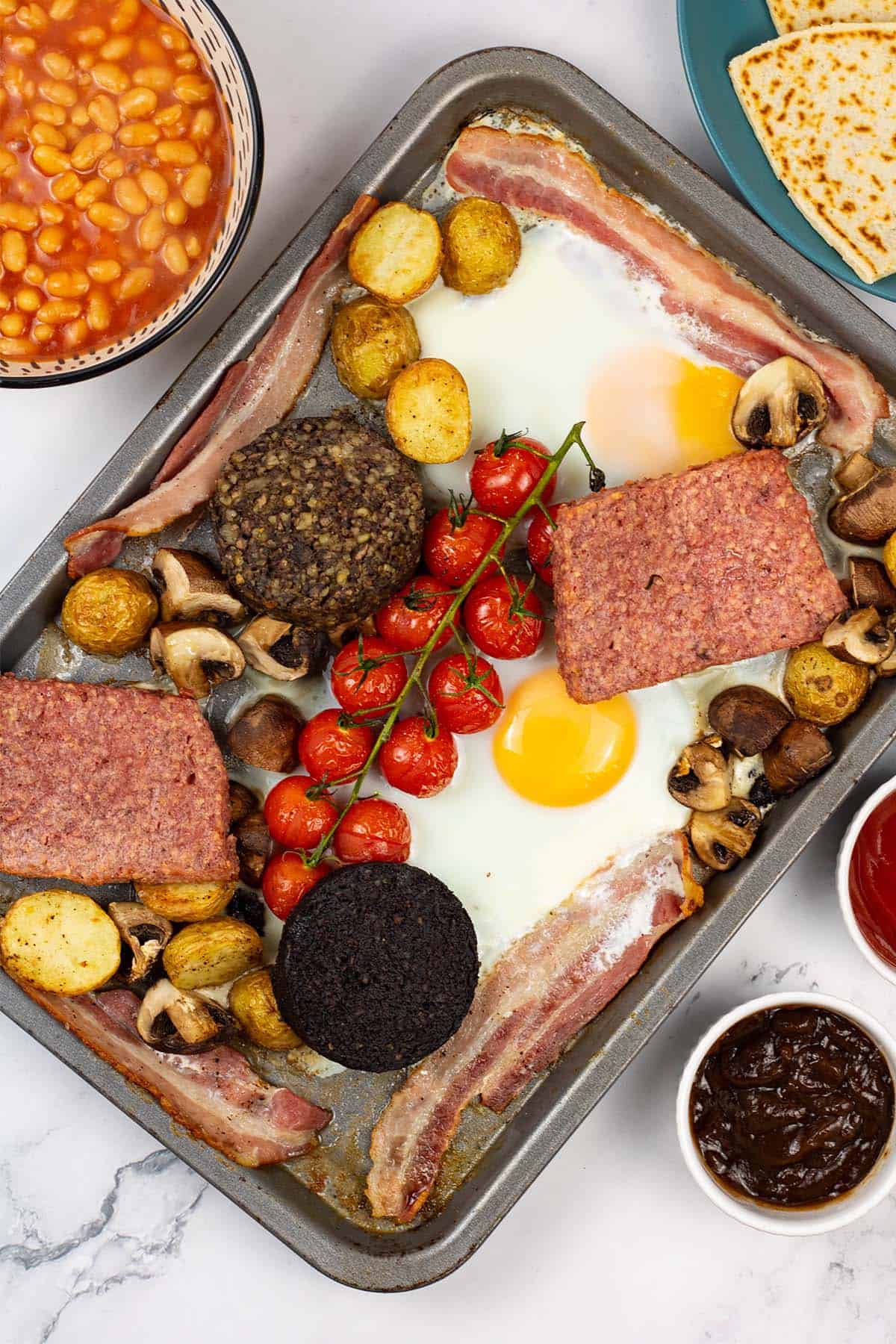Scottish breakfast traybake on a baking tray with a bowl of baked beans, plate of tattie scones and small dishes of brown and tomato sauces on the side.
