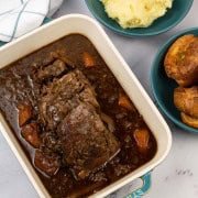 Slow cooked brisket with red wine, thyme and onions in a rectangular serving dish with bowls of mashed potatoes and Yorkshire puddings on the side.