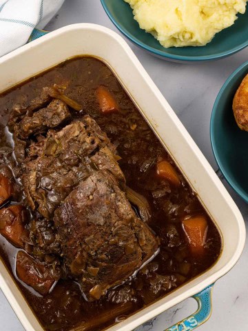Slow cooked brisket with red wine, thyme and onions in a rectangular serving dish with bowls of mashed potatoes and Yorkshire puddings on the side.