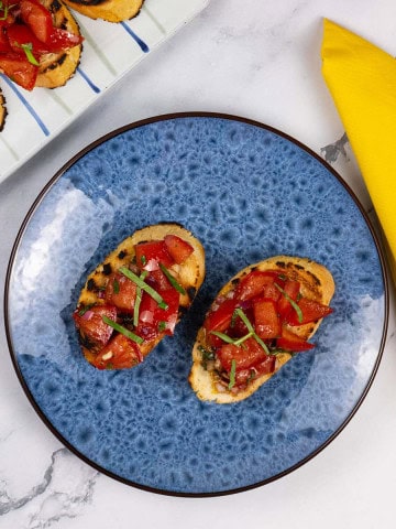 A portion of tomato bruschetta on a blue side plate with a striped platter of more bruschetta on the side.