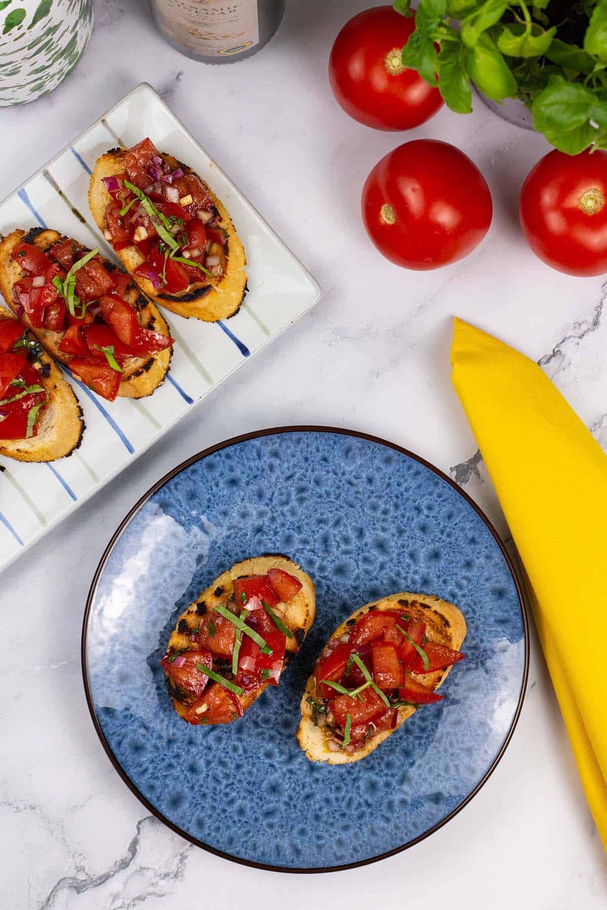 A portion of tomato bruschetta on a blue side plate with a striped platter of more bruschetta on the side.