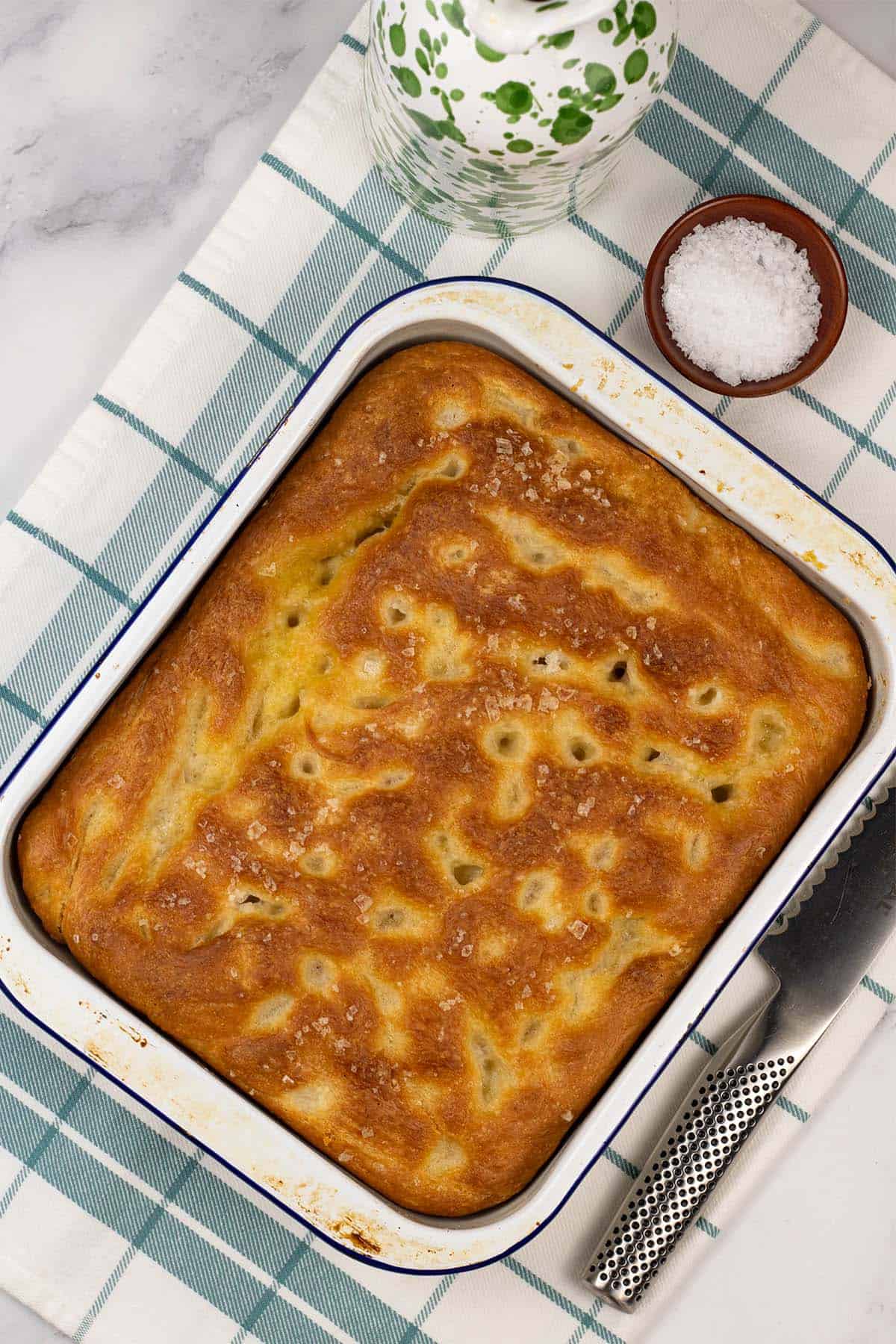 Authentic focaccia in a white and blue rimmed enamel dish, sitting on a teal and white checked towel.