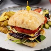 Halloumi burger on a white and black dinner plate with chunky oven chips and greek salad.