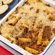 Beef and tomato pasta bake in a blue rimmed white enamel roasting tin with a plate of sliced crusty bread in the background.