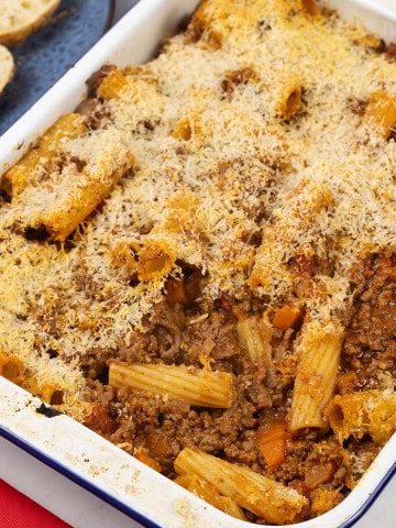 Beef and tomato pasta bake in a blue rimmed white enamel roasting tin with a plate of sliced crusty bread in the background.