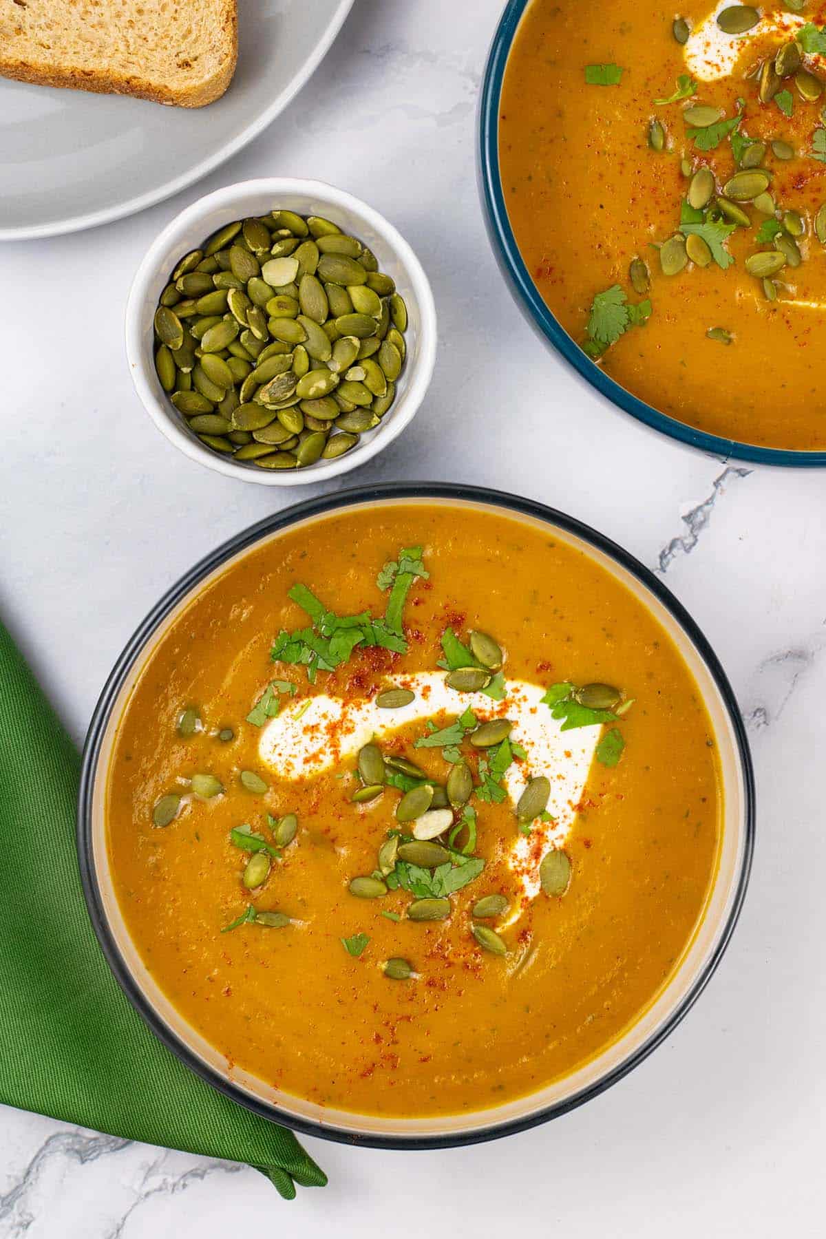 Two bowls of carrot and coriander soup with a small dish of toasted pumpkin seeds and plate of bread in the background.