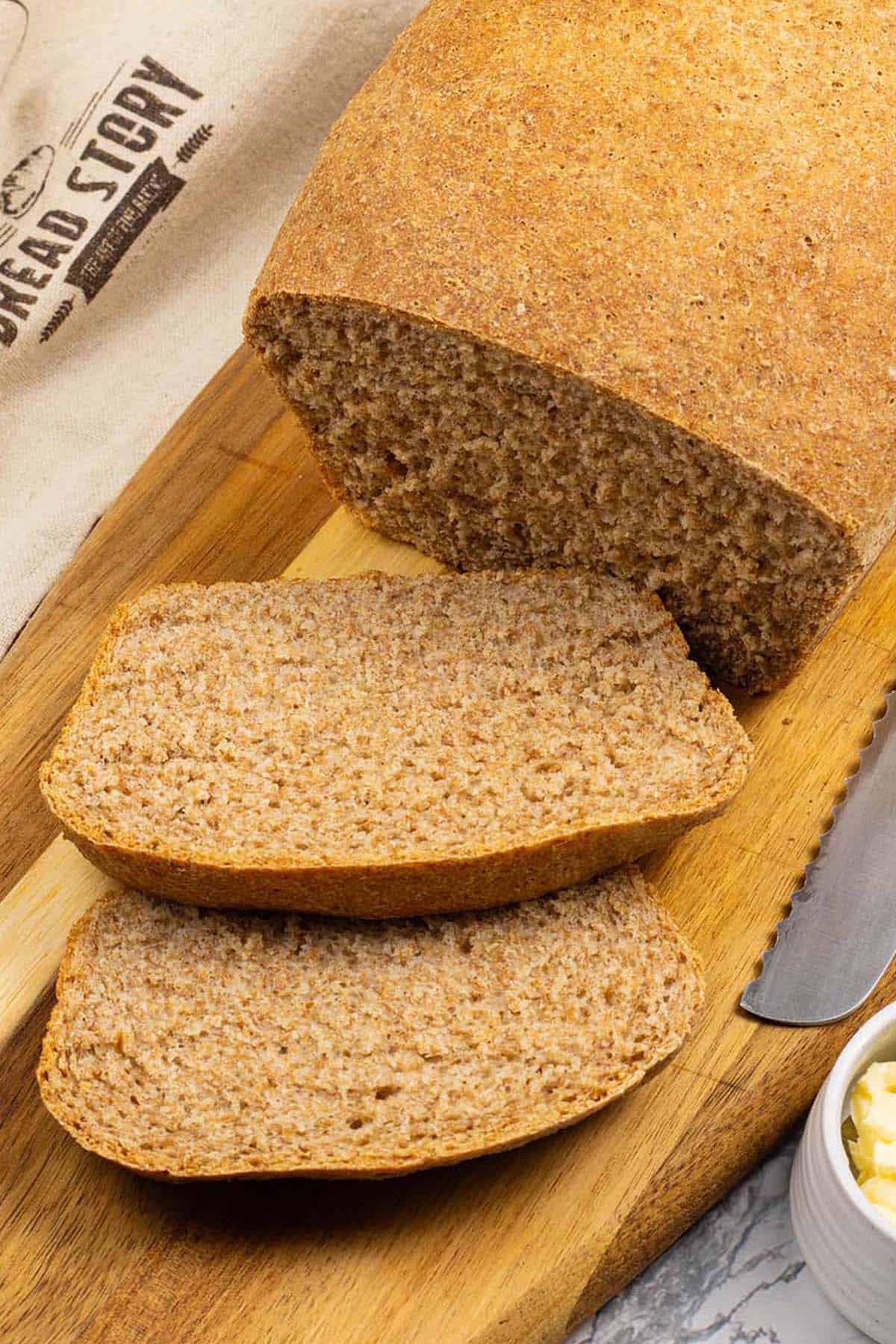 Classic wholemeal bread made in the Thermomix on a board with 2 slices cut and a small dish of butter with butter knife on the side.