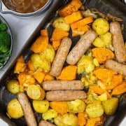 Sausage, squash and potato bake in a large roasting tin with a jug of onion gravy and bowl of broccoli and green beans on the side.