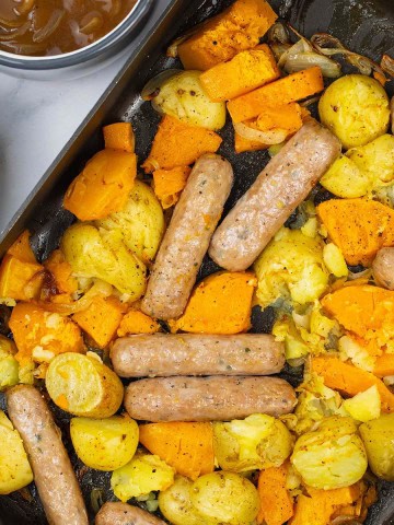 Sausage, squash and potato bake in a large roasting tin with a jug of onion gravy and bowl of broccoli and green beans on the side.