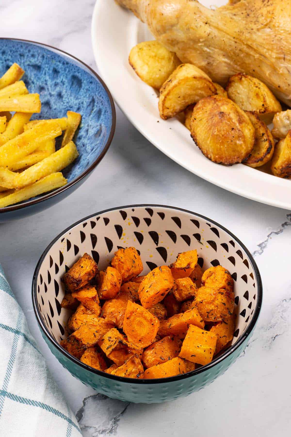 Air fryer carrots and butternut squash in a teal, black and cream patterned bowl with a bowl of parsnips and platter of roast chicken and potatoes in the background.