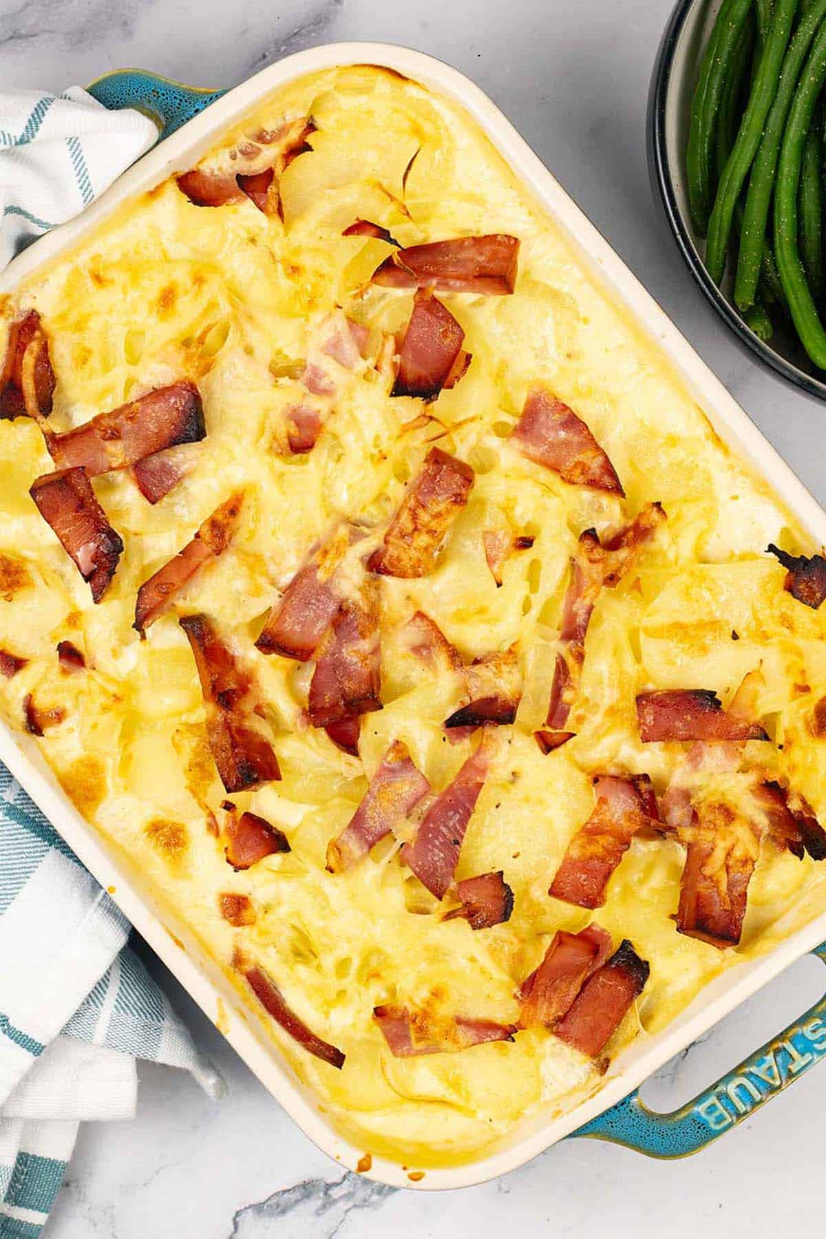 Cheesy potato and gammon bake in a rectangular Staub oven dish with a bowl of green beans and broccoli on the side.