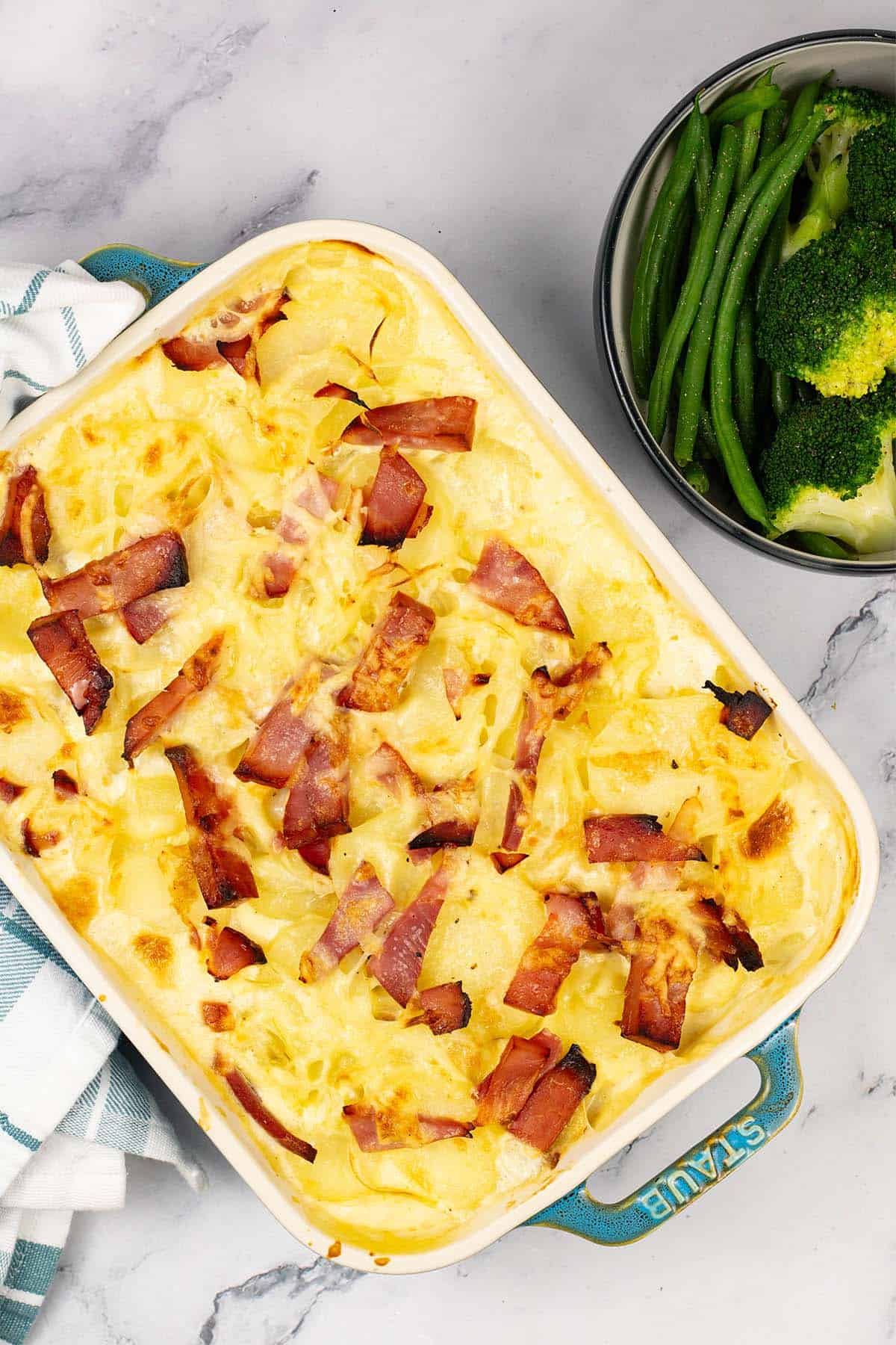 Cheesy potato and gammon bake in a rectangular Staub oven dish with a bowl of green beans and broccoli on the side.