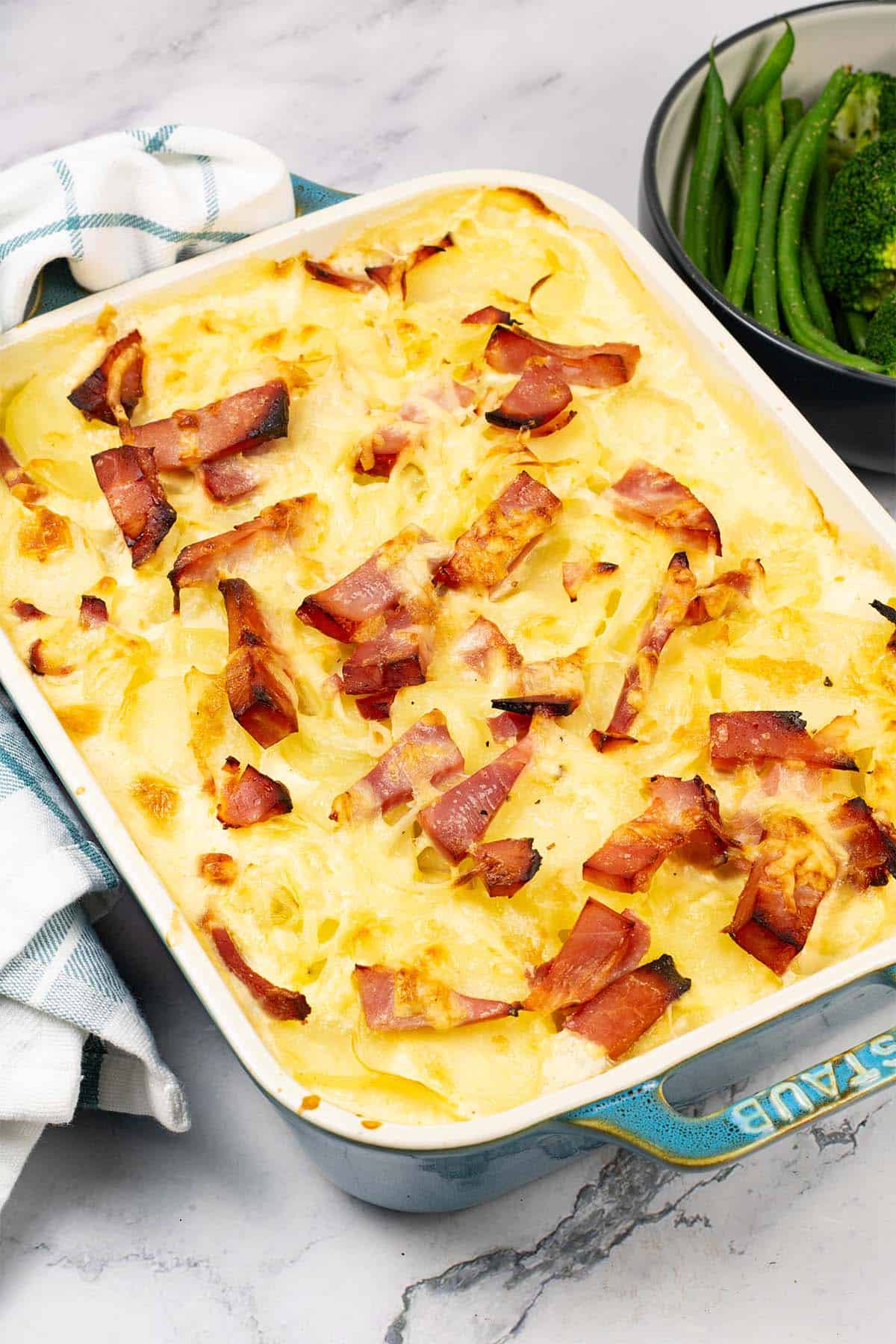 Cheesy potato and gammon bake in a rectangular Staub oven dish with a bowl of green beans and broccoli on the side.