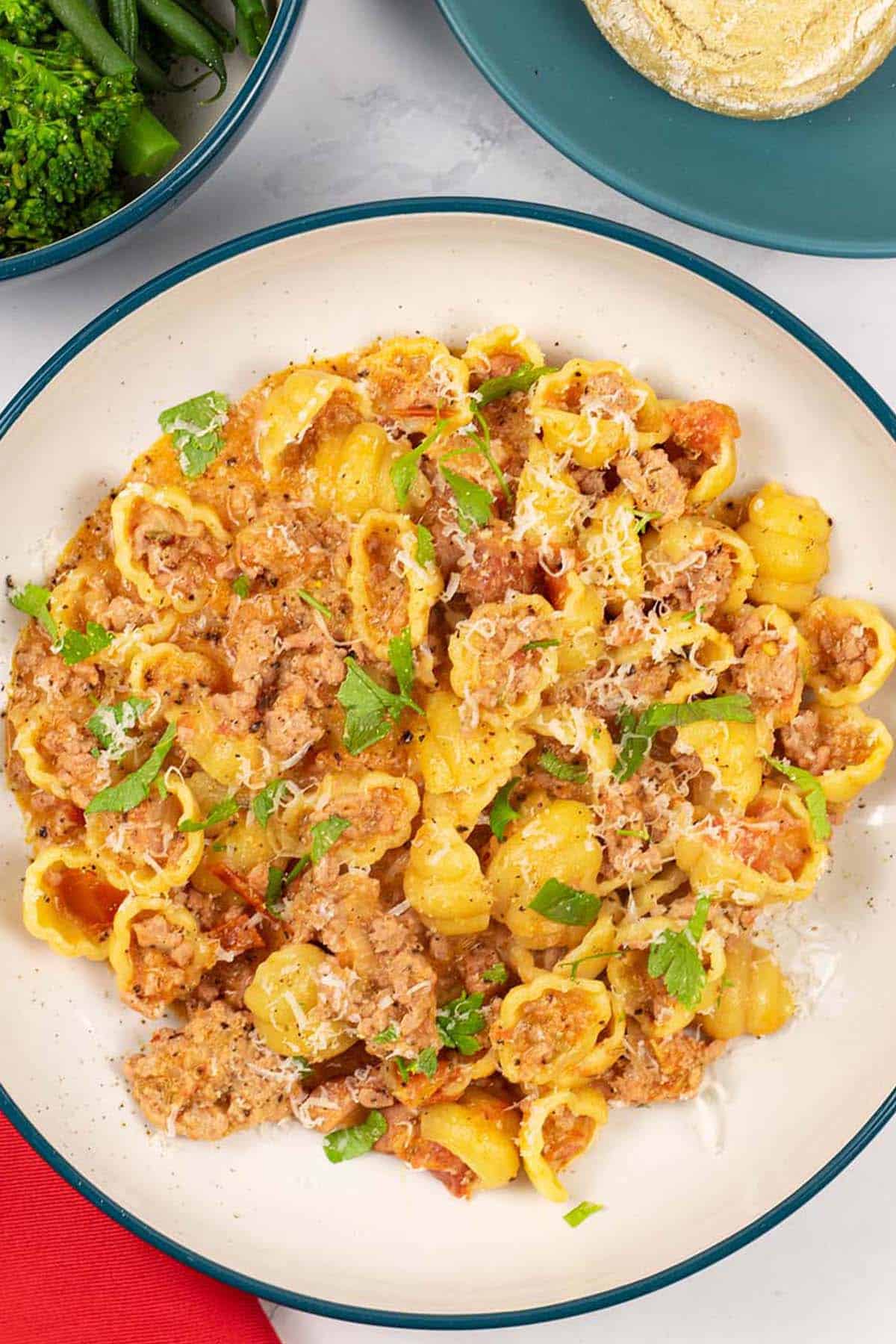 A portion of creamy Italian pork pasta in a teal and cream bowl with a bowl of broccoli and plate of crusty buns on the side.