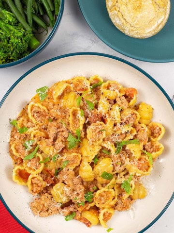 A portion of creamy Italian pork pasta in a teal and cream bowl with a bowl of broccoli and plate of crusty buns on the side.