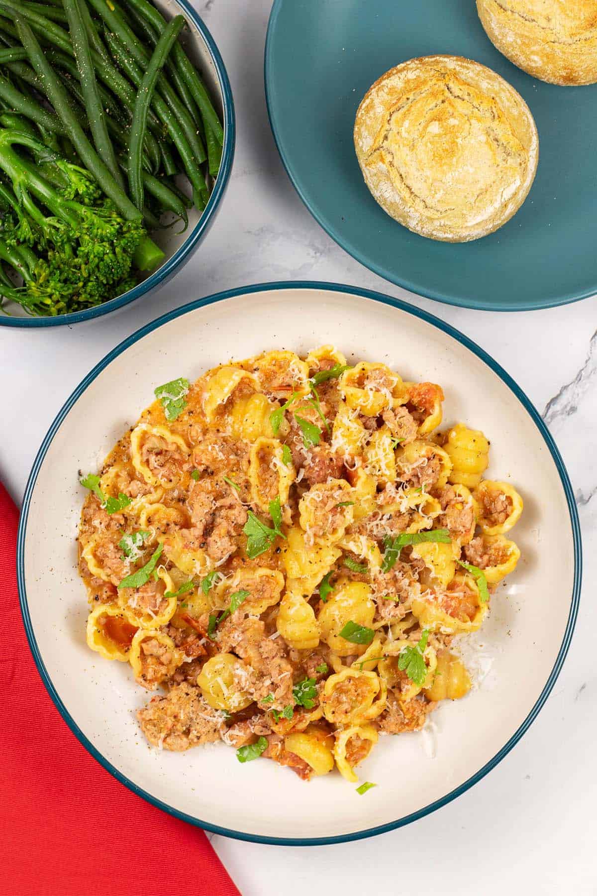 A portion of creamy Italian pork pasta in a teal and cream bowl with a bowl of broccoli and plate of crusty buns on the side.