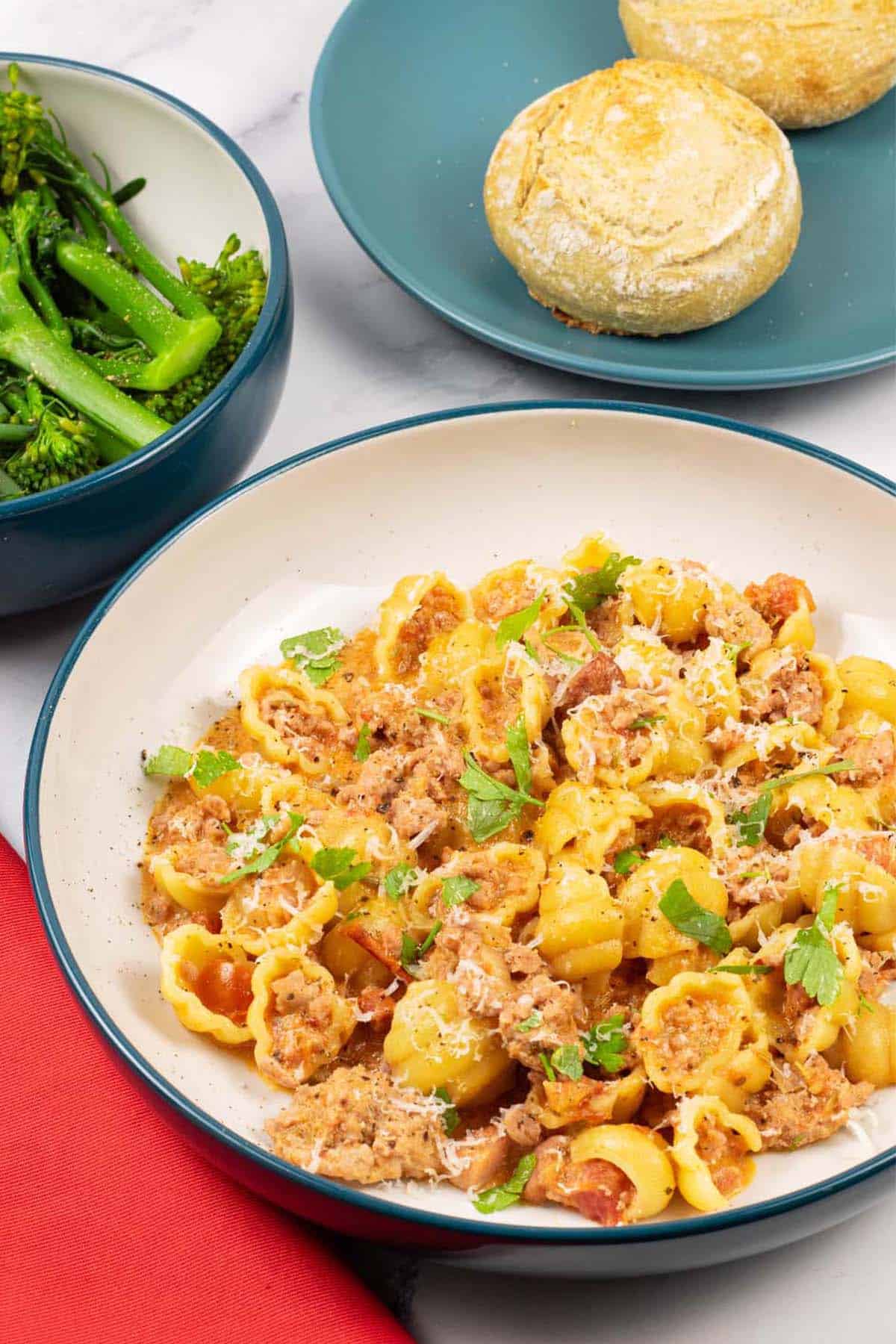 A portion of creamy Italian pork pasta in a teal and cream bowl with a bowl of broccoli and plate of crusty buns on the side.