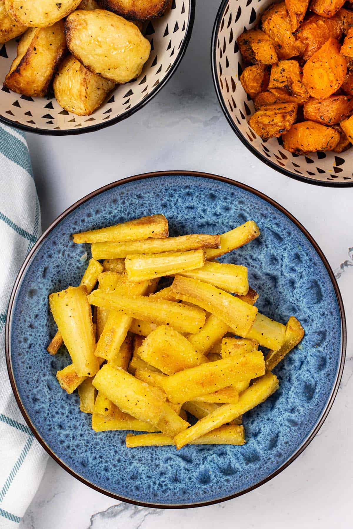 Crisp honey mustard parsnips in a blue patterned serving bowl with bowls of roast potatoes and air fryer carrots and butternut squash in the background.