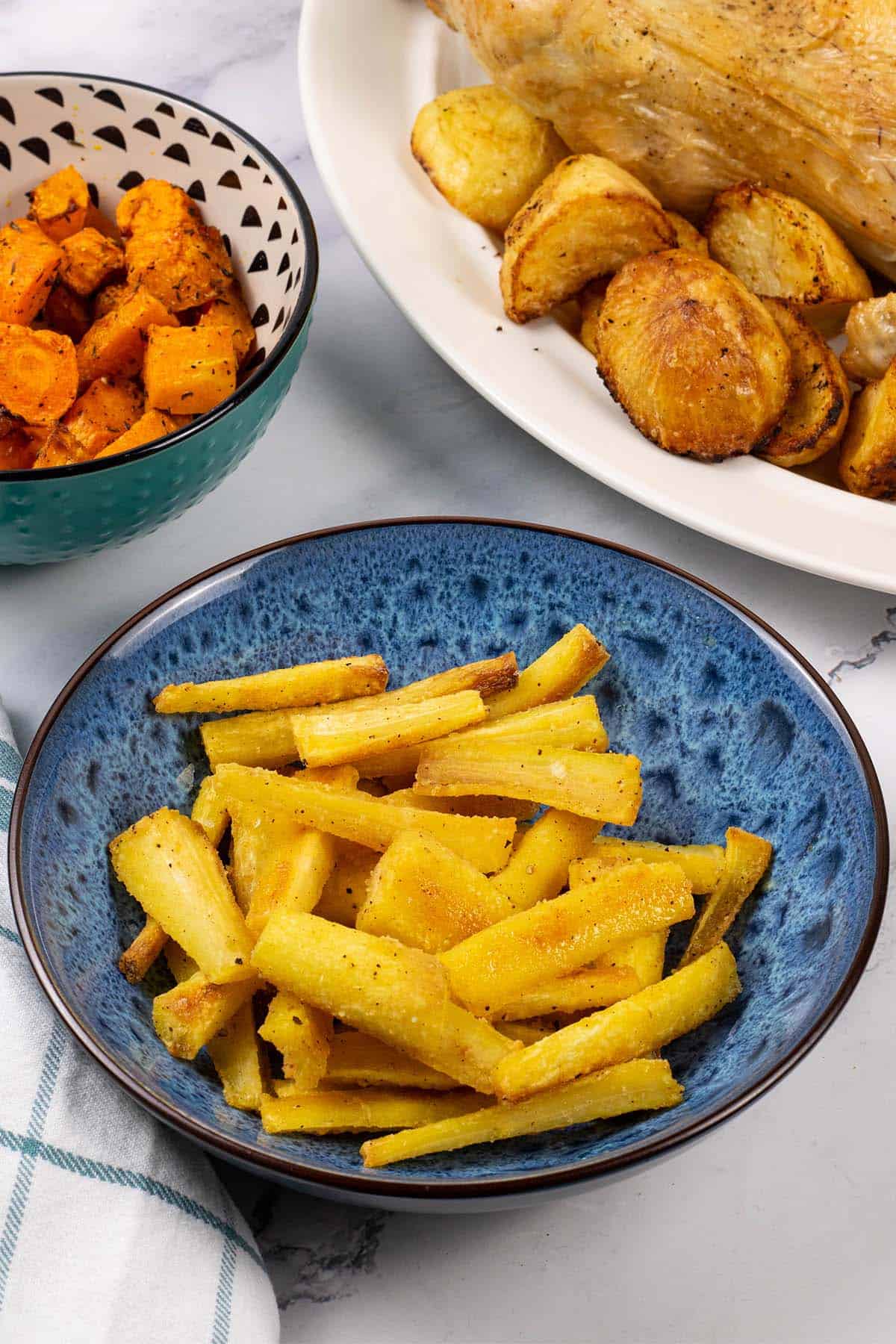 Crisp honey mustard parsnips in a blue patterned serving bowl with a roast chicken, potatoes and air fryer carrots and butternut squash in the background.