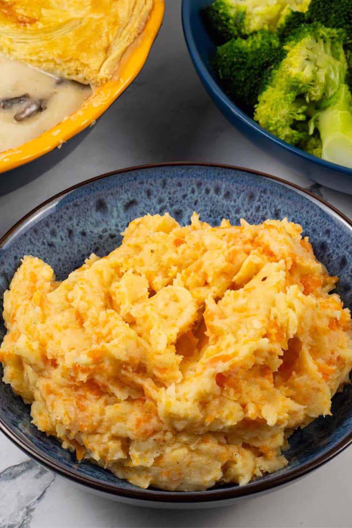 Root veg mash in a blue patterned serving bowl with a pie and bowl of broccoli and green beans in the background.