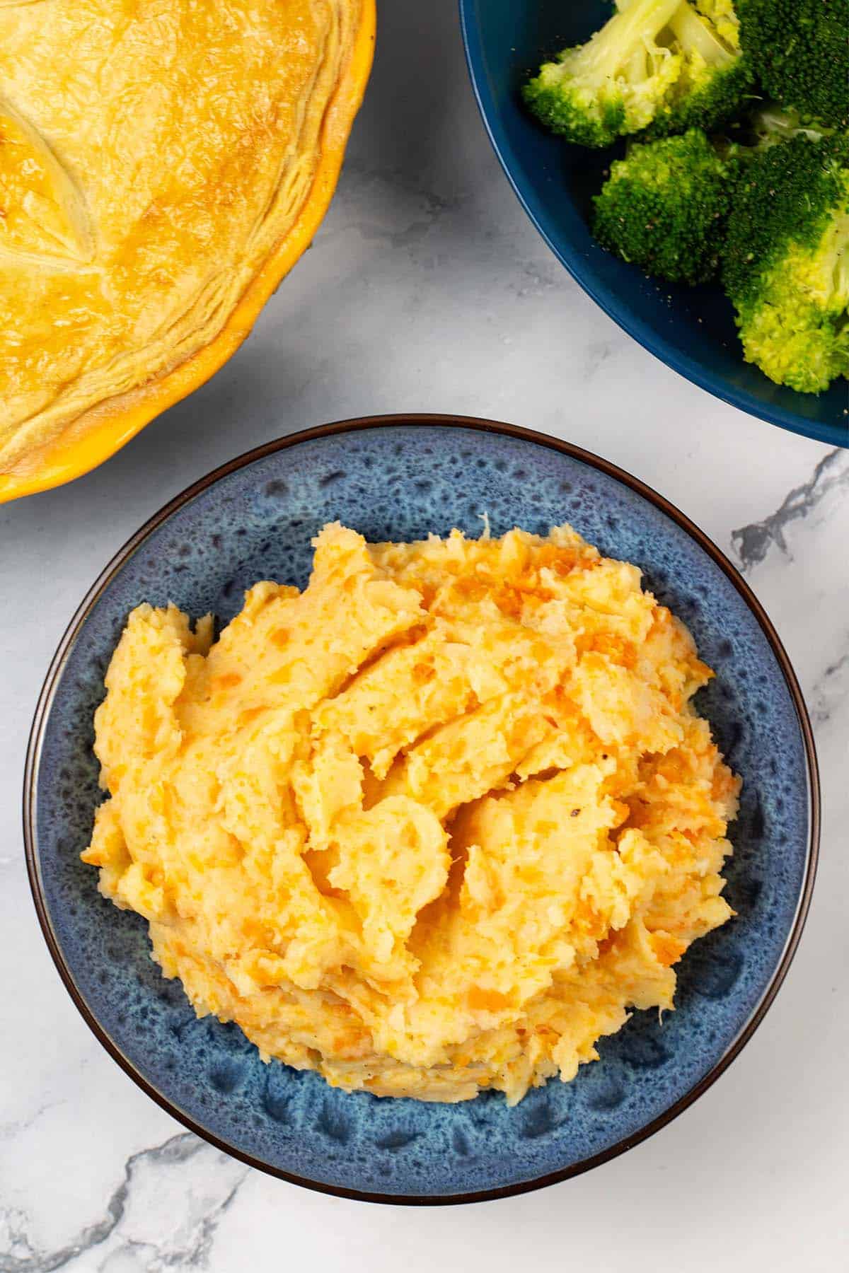 Root veg mash in a blue patterned serving bowl with a pie and bowl of broccoli and green beans in the background.
