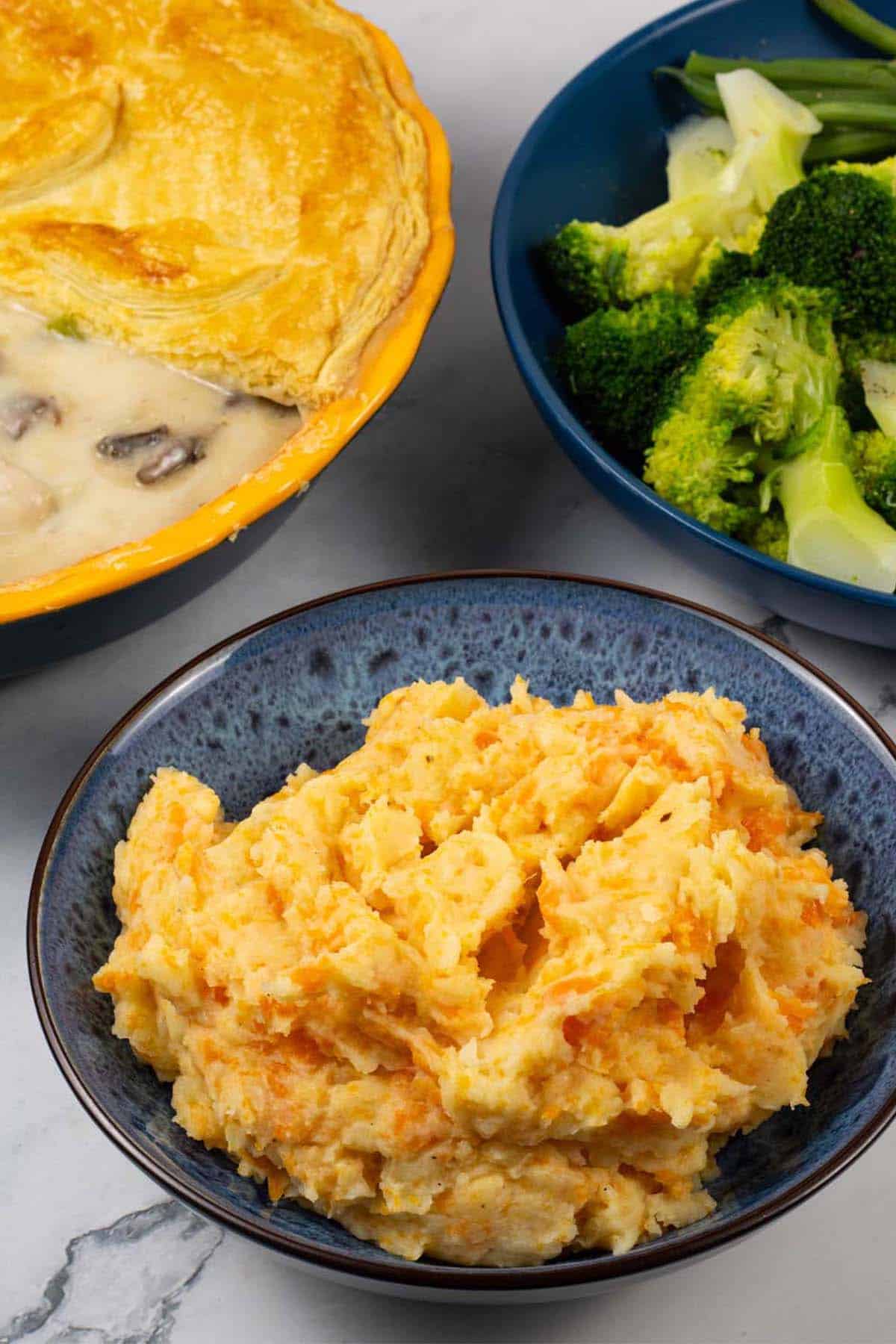 Root veg mash in a blue patterned serving bowl with a pie and bowl of broccoli and green beans in the background.