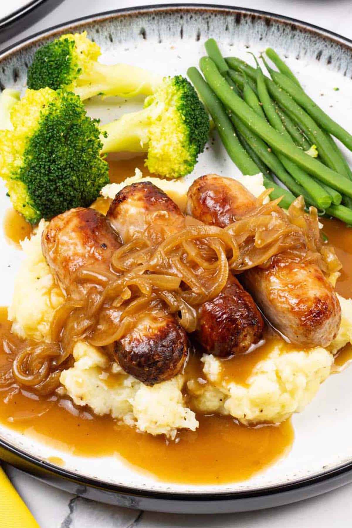 A plate of sausages with caramelised onion gravy on a bed of mashed potatoes with broccoli and green beans on the side.