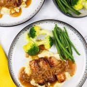 Two plates of sausages with caramelised onion gravy on a bed of mashed potatoes with broccoli and green beans on the side.