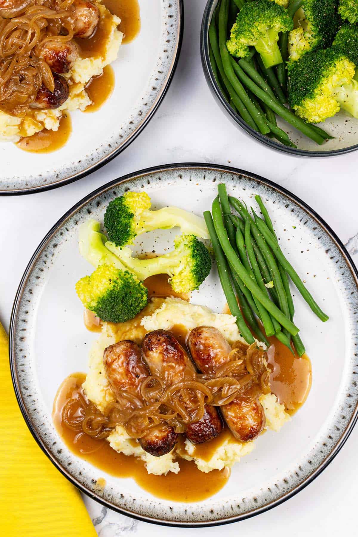 Two plates of sausages with caramelised onion gravy on a bed of mashed potatoes with broccoli and green beans on the side.
