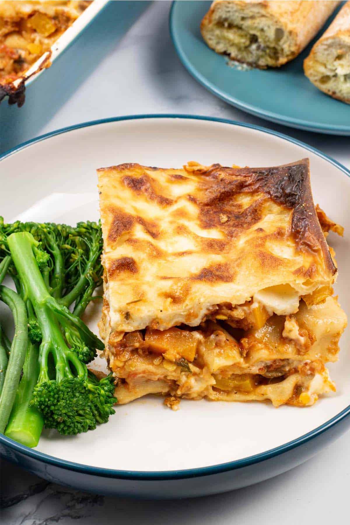 A portion of vegetable lasagne in a teal and cream bowl with tenderstem broccoli and green beans on the side.