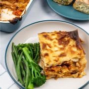 A portion of vegetable lasagne in a teal and cream bowl with tenderstem broccoli and green beans on the side.