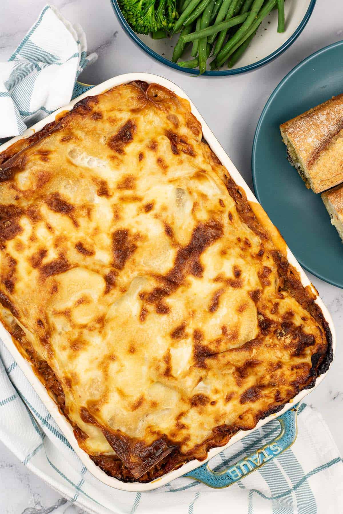 Vegetable lasagne in a Staub ovenproof dish with a bowl of broccoli and green beans, and plate of garlic baguette on the side.