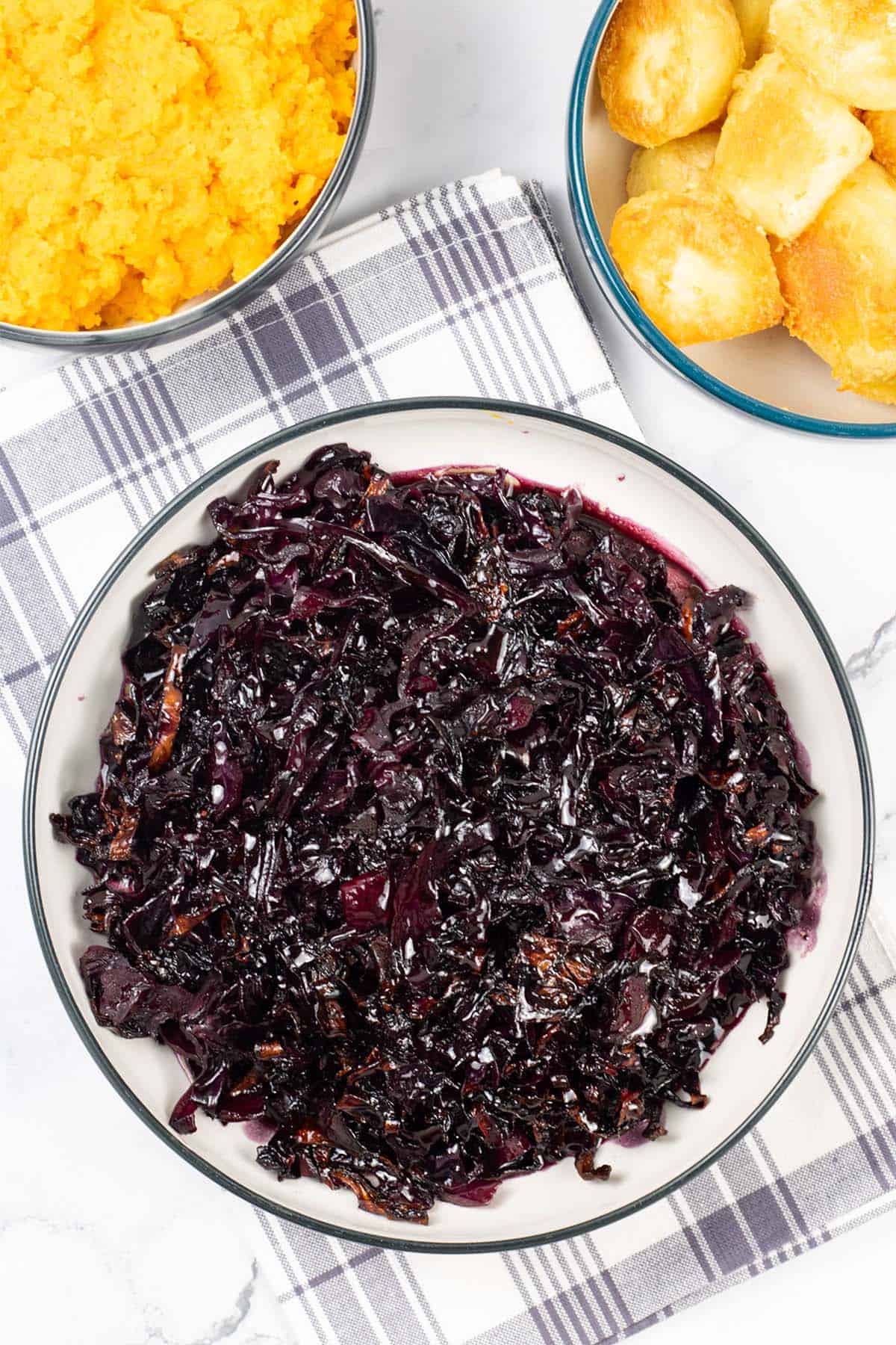 Braised red cabbage in a black and cream serving bowl with bowls of roast potatoes and carrot and swede mash in the background.