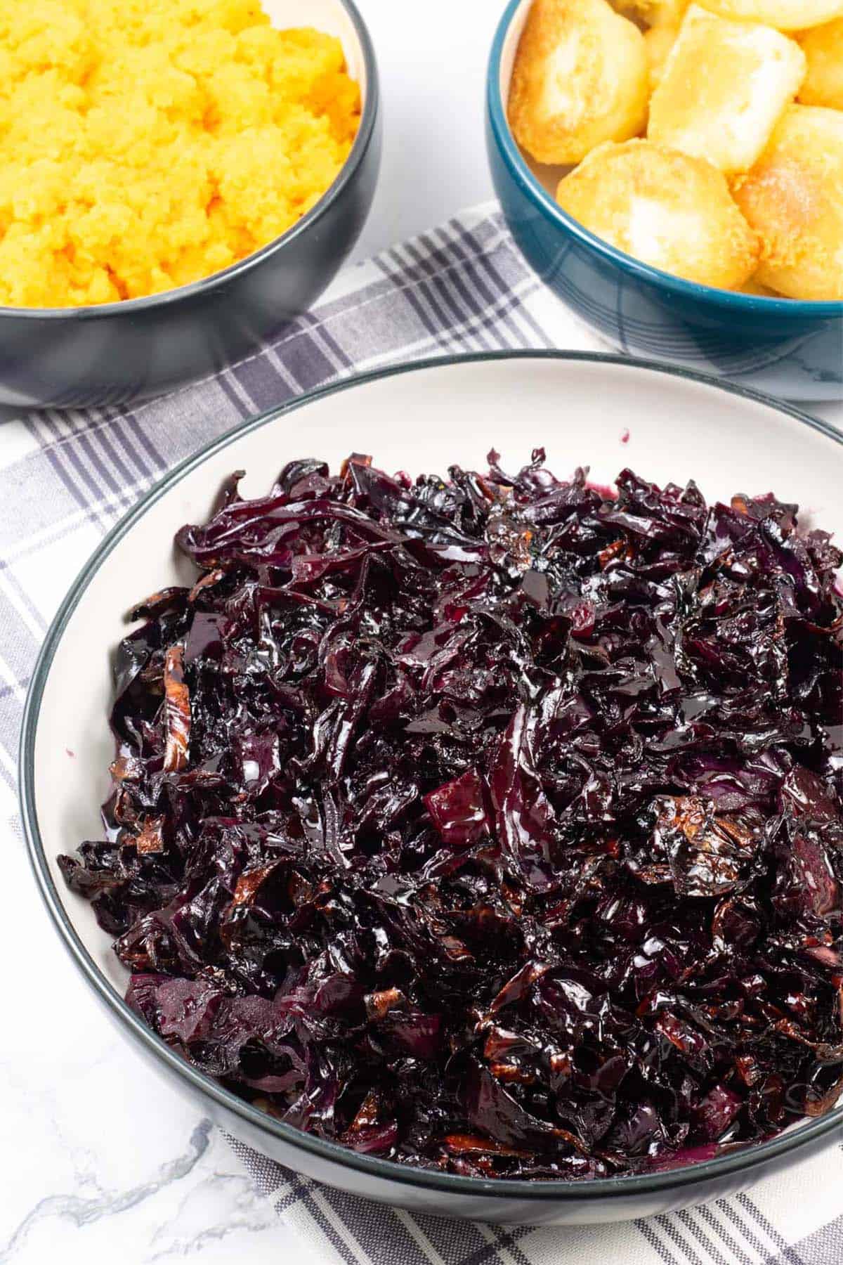 Braised red cabbage in a black and cream serving bowl with bowls of roast potatoes and carrot and swede mash in the background.