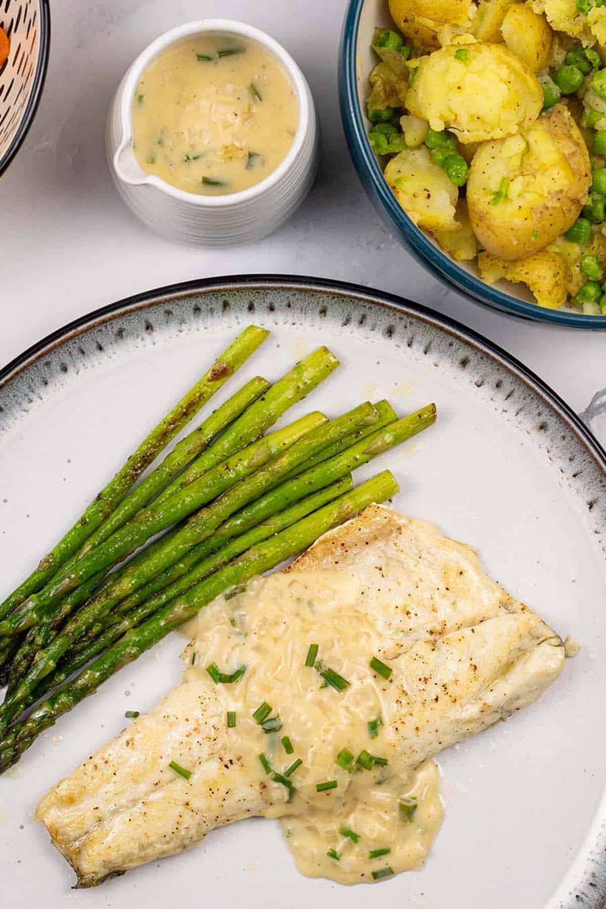 A portion of easy sea bass with white wine and cream sauce on a black and light grey patterned dinner plate with sautéed asparagus.