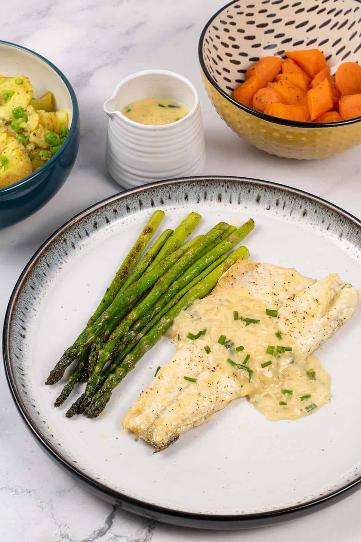 A portion of easy sea bass with white wine and cream sauce on a black and light grey patterned dinner plate with sautéed asparagus.