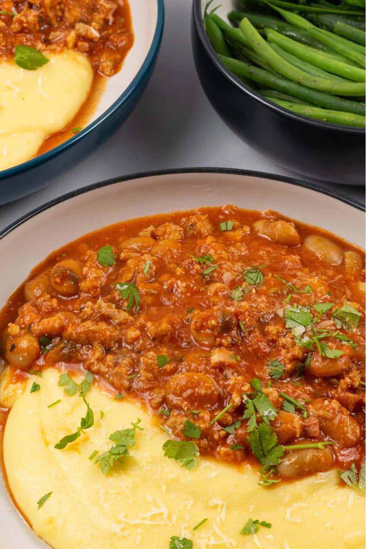 Close up of two bowls of sausage and white bean ragu with cheesy polenta and a bowl of green beans and broccoli on the side.