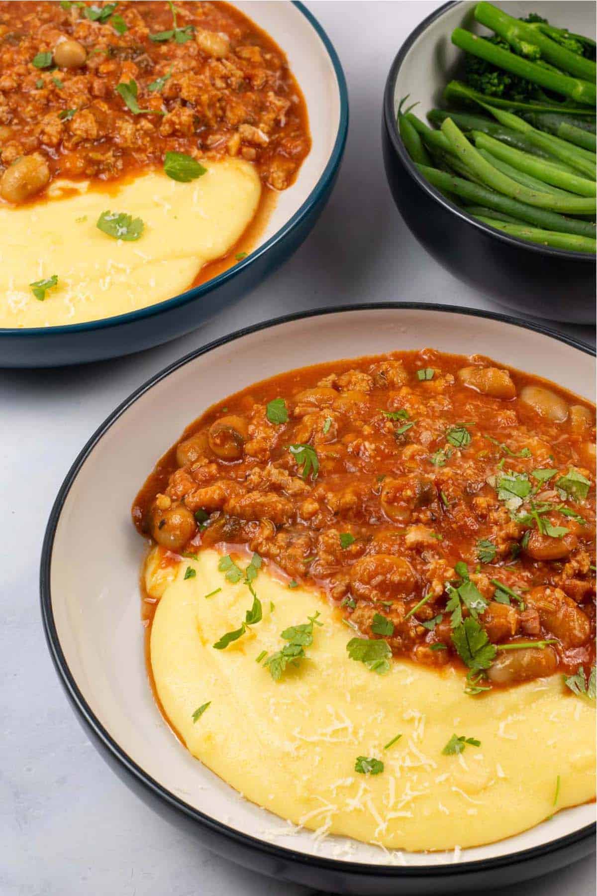 Two bowls of sausage and white bean ragu with cheesy polenta and a bowl of green beans and broccoli on the side.