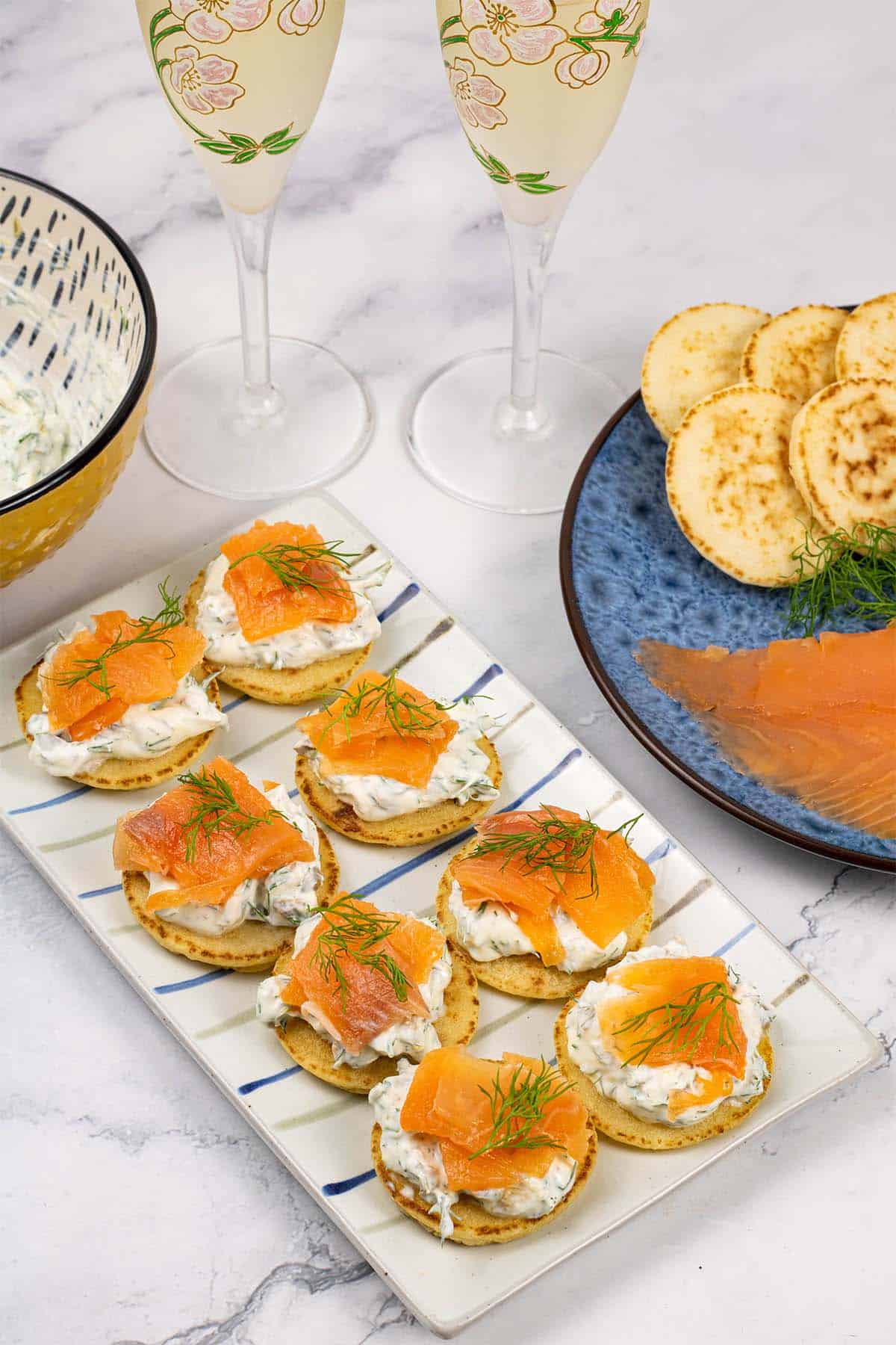 8 smoked salmon and lemon caper cream cheese blinis on a rectangle striped platter with 2 glasses of champagne in the background.