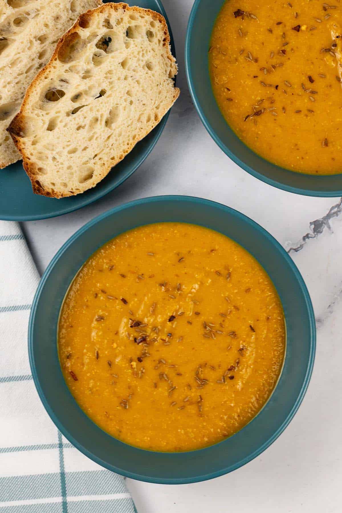 Two bowls of spiced carrot and lentil soup with a plate of sliced crusty bread on the side.