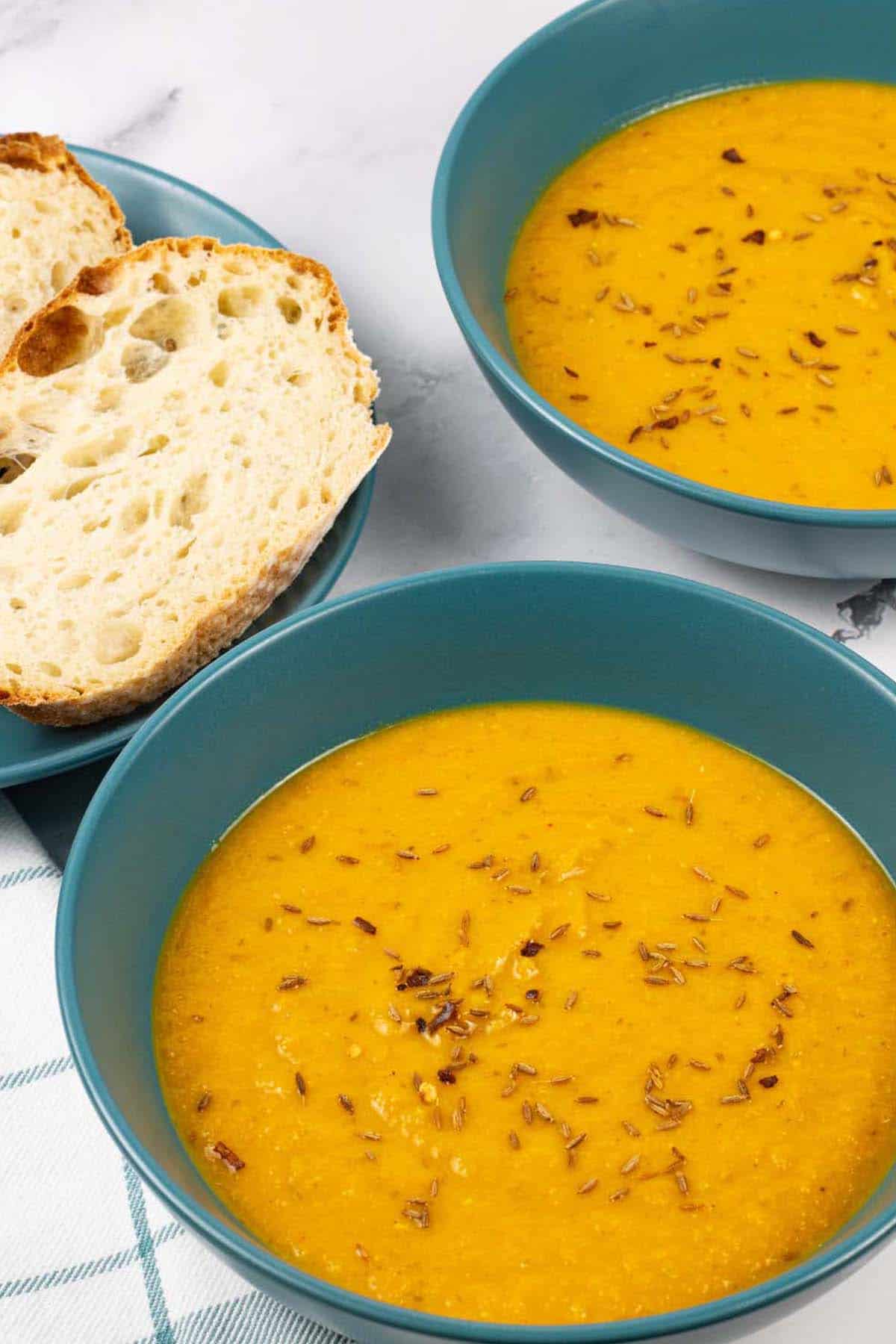 Two bowls of spiced carrot and lentil soup with a plate of sliced crusty bread on the side.
