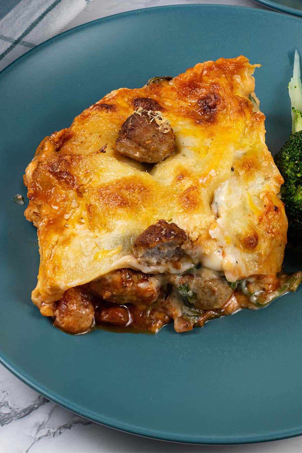 Close up of a portion of three cheese meatball lasagne on a teal dinner plate with broccoli.